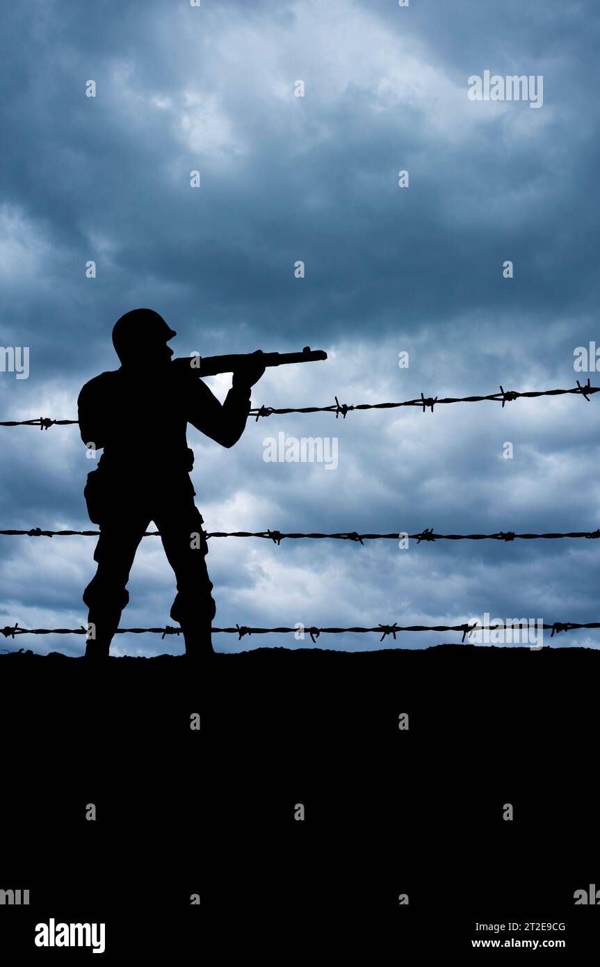 silhouette di un soldato che punta una pistola dietro un filo spinato Foto Stock