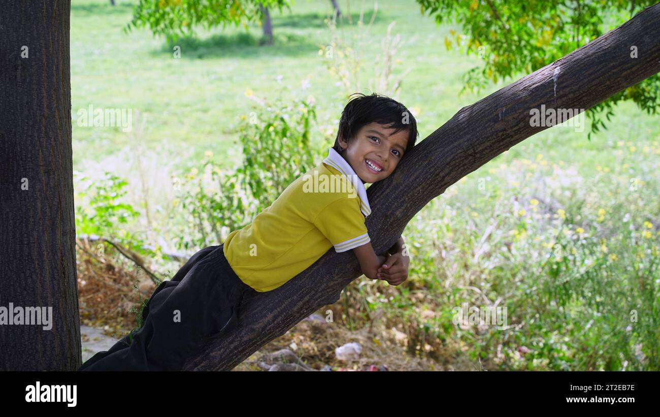 Felice ragazzo sorridente appeso ad un albero. Il bambino ride felicemente. Atmosfera primaverile. Stile di vita attivo. Arrampicata sugli alberi. Foto Stock