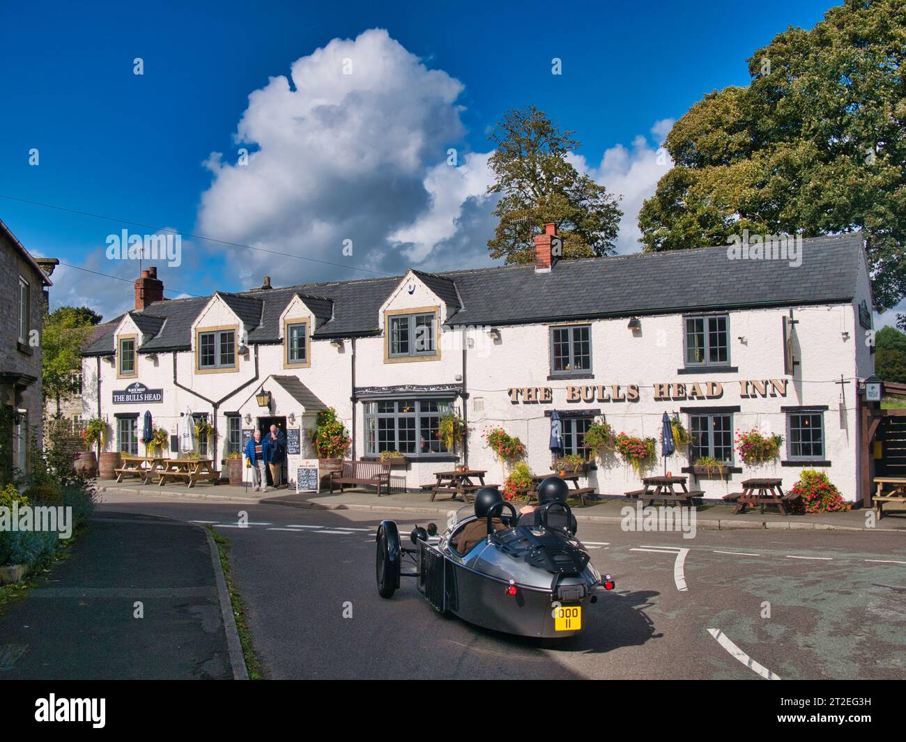 Un'auto sportiva Morgan a tre ruote color argento si ferma all'incrocio di fronte al Bulls Head Inn a Foolow, Derbyshire, Regno Unito. Preso in una giornata di sole. Foto Stock