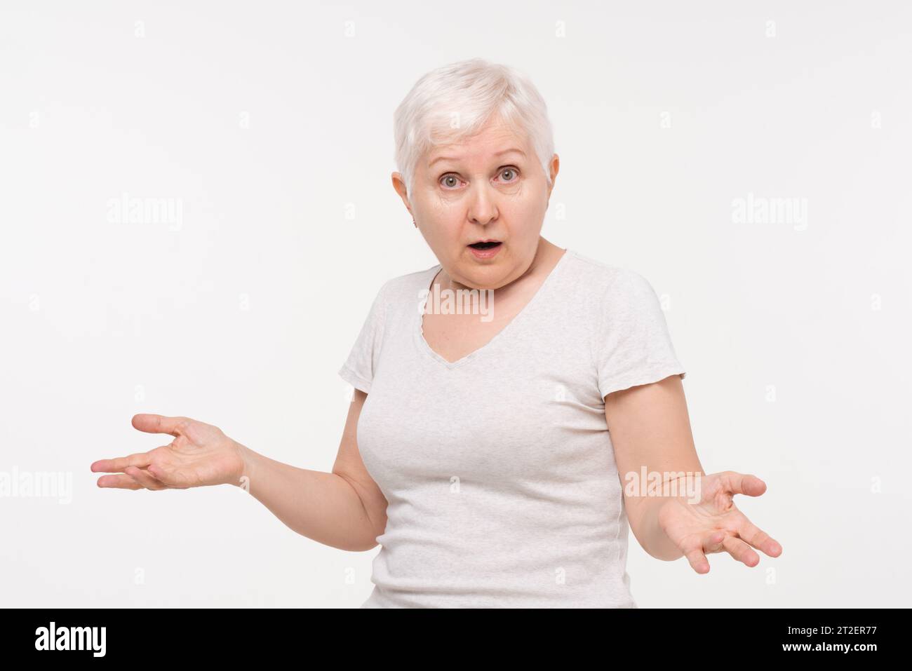 Anziana donna caucasica anziana che esprime una sensazione scioccata e confusa con il gesto della mano aperta, ripresa in studio su sfondo bianco isolato Foto Stock