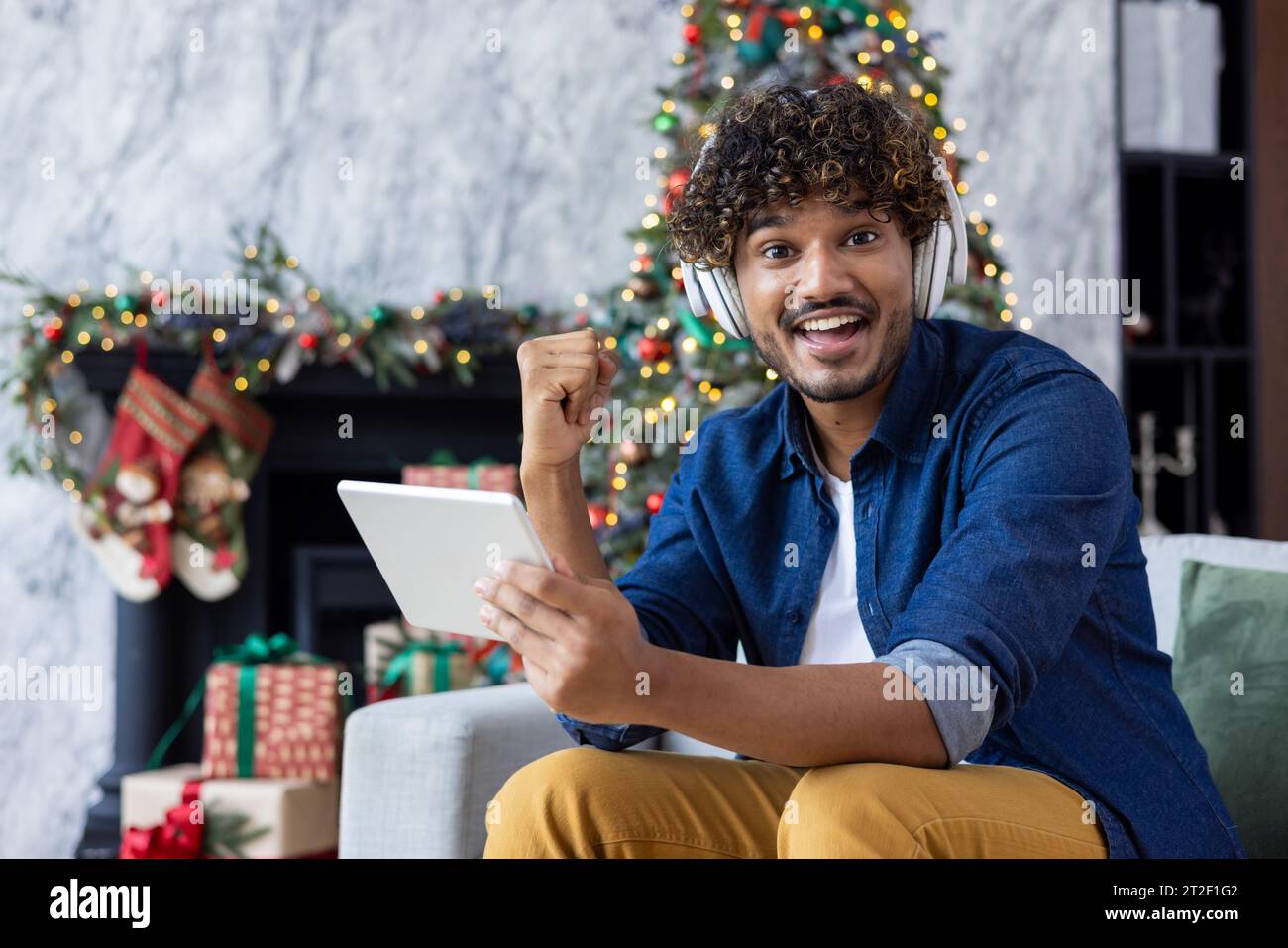 Ritratto di un giovane indiano felice che è felice davanti alla telecamera che mostra un gesto di vittoria con la mano alla fotocamera. Seduto a casa durante le vacanze di Natale e tenendo in mano un tablet. Foto Stock