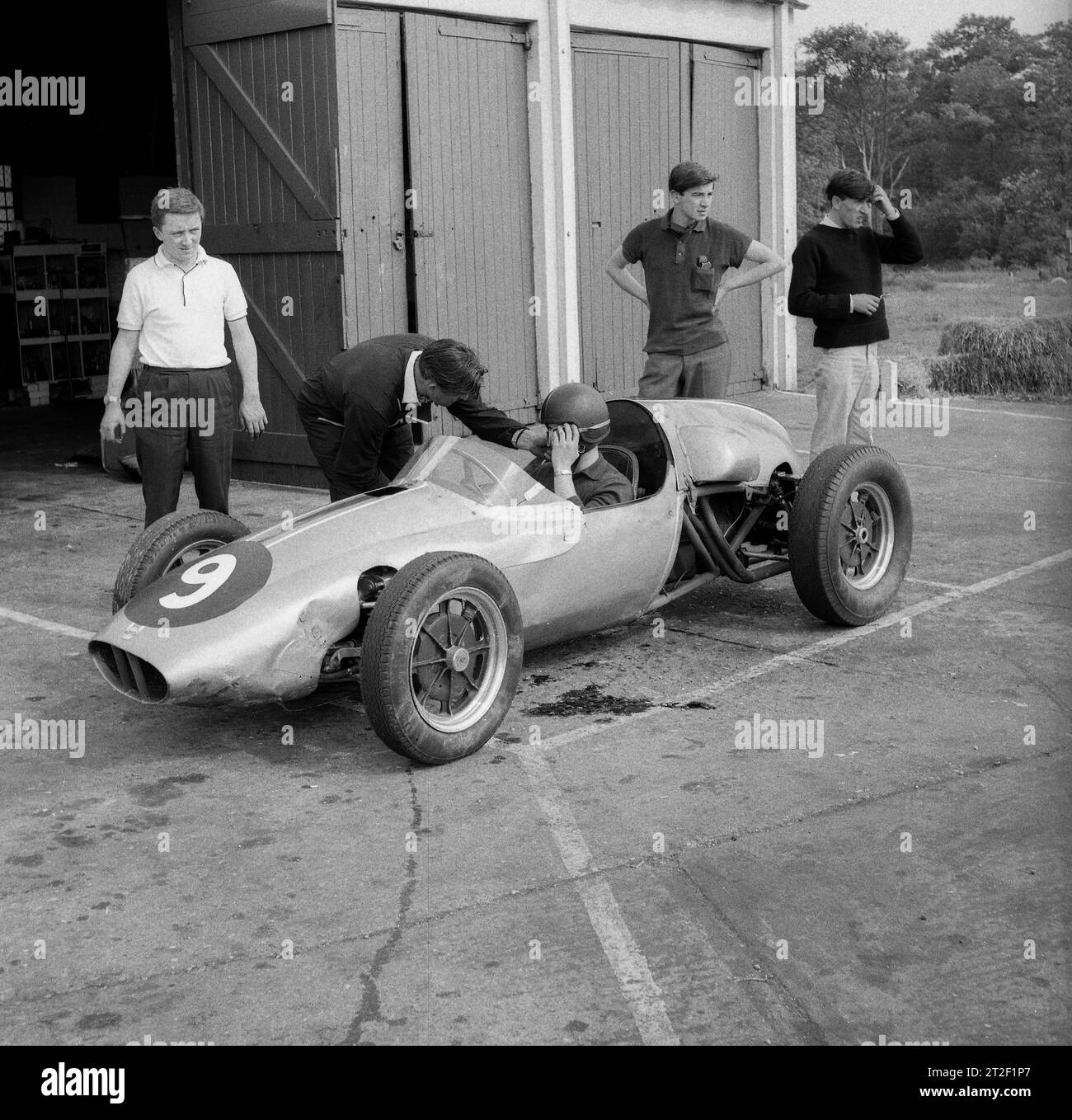 1962, storico, un giovane pilota maschio seduto su un'auto da corsa Cooper aperta che riceve istruzioni all'esterno di un capannone presso l'aeroporto di Finmere, Finemere, Bucks, Inghilterra, Regno Unito, a casa in questo periodo della Geoff Clarkes Motor School. La vettura da corsa Cooper, con il suo motore dietro il pilota, era innovativa all'epoca e divenne la prima vettura con motore posteriore a vincere una gara di Formula 1. Foto Stock