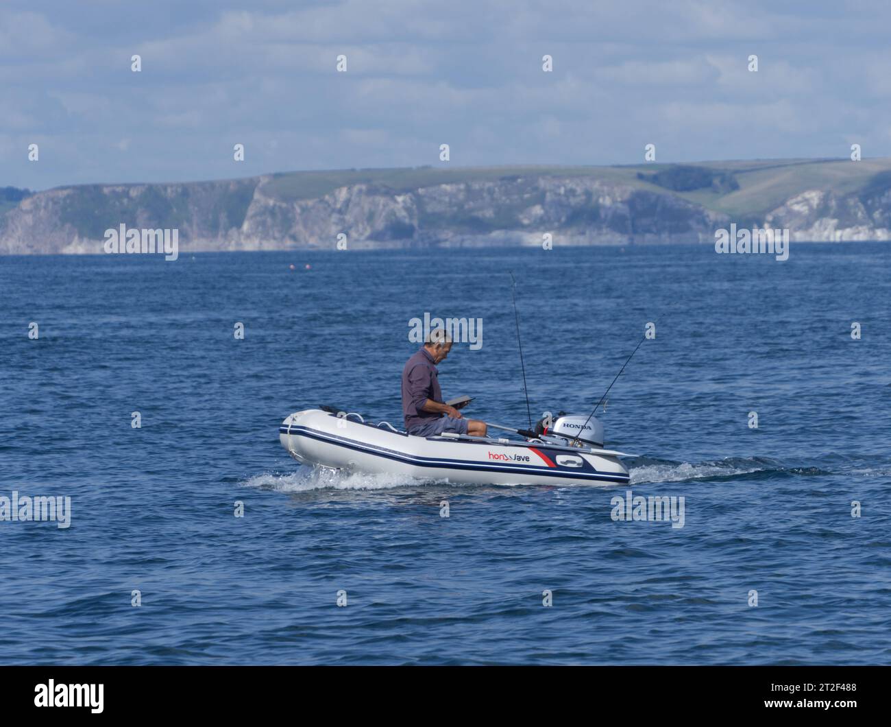 Pescatore libero con tablet/ipad mentre pesca in mare da una piccola barca con Bigbury Bay sullo sfondo Foto Stock