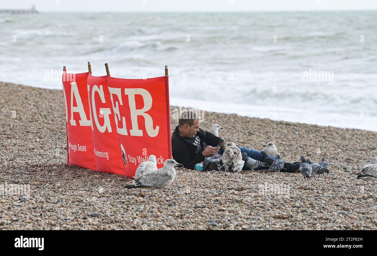 Brighton Regno Unito 19 ottobre 2023 - i visitatori di fronte a un cartello di pericolo sulla spiaggia di Brighton in una giornata ventosa lungo la costa meridionale mentre Storm Babet colpisce parti della Gran Bretagna con avvisi meteo rossi emessi in Scozia: Credit Simon Dack / Alamy Live News Foto Stock