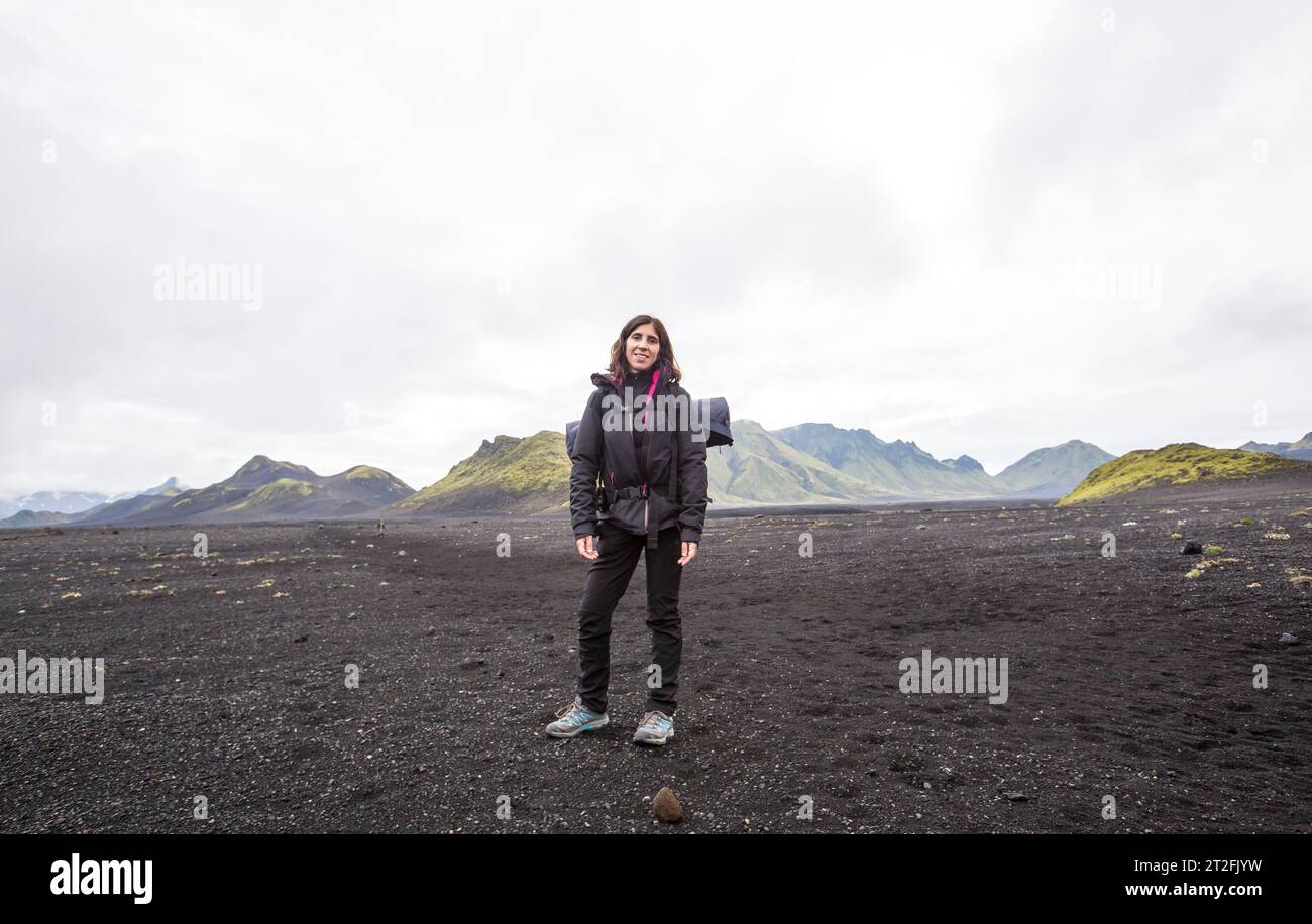 Una giovane donna in cenere vulcanica e una montagna verde sullo sfondo durante il trekking di 54 km da Landmannalaugar, Islanda Foto Stock