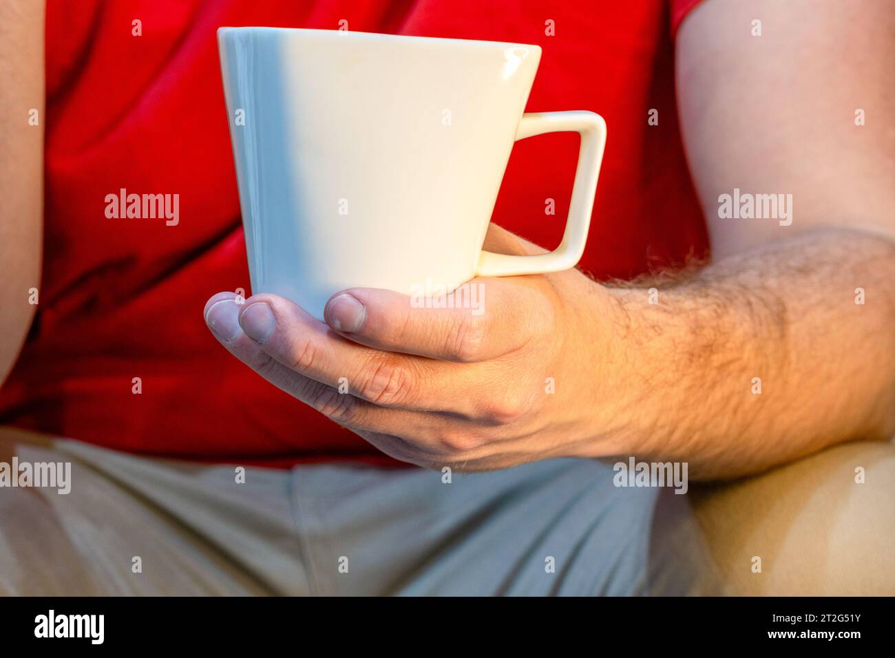 Uomo latino caucasico, seduto, vestito con una t-shirt rossa e jeans. Tenendo una tazza bianca. Foto Stock