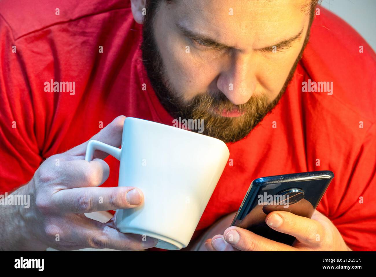 Uomo latino caucasico, seduto, vestito con una t-shirt rossa e jeans. Tenendo in mano un cellulare e una tazza bianca. Foto Stock