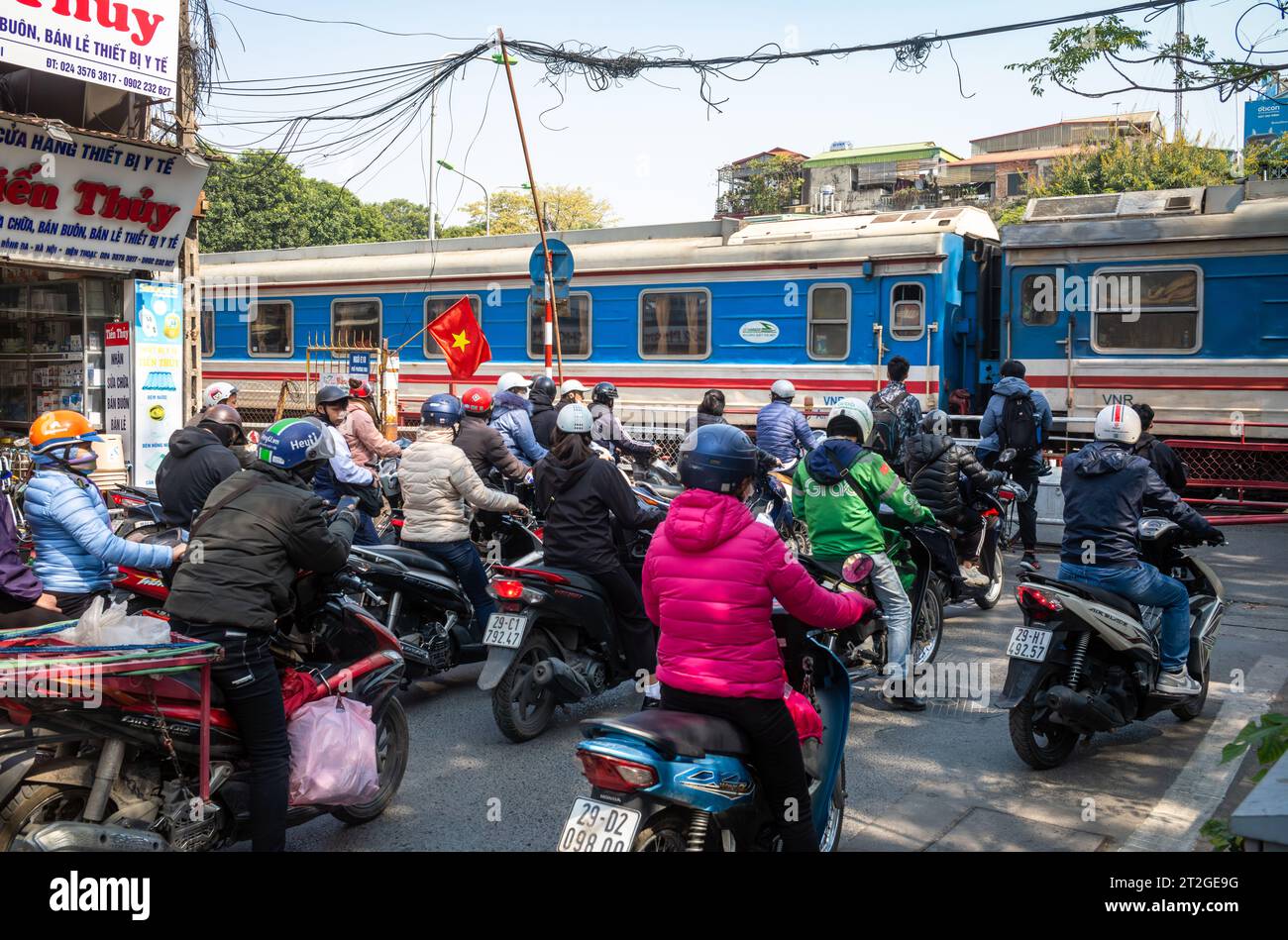Un treno della Vietnam Railways passa le persone in moto e biciclette ad un passaggio a livello (attraversamento ferroviario) ad Hanoi, in Vietnam. Foto Stock