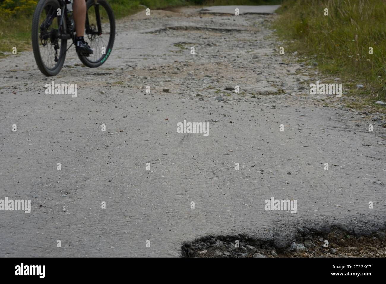 Vecchia strada asfaltata con molte buche e il ciclista che cerca di evitare gli ostacoli che corrono sul lato della strada. Foto Stock