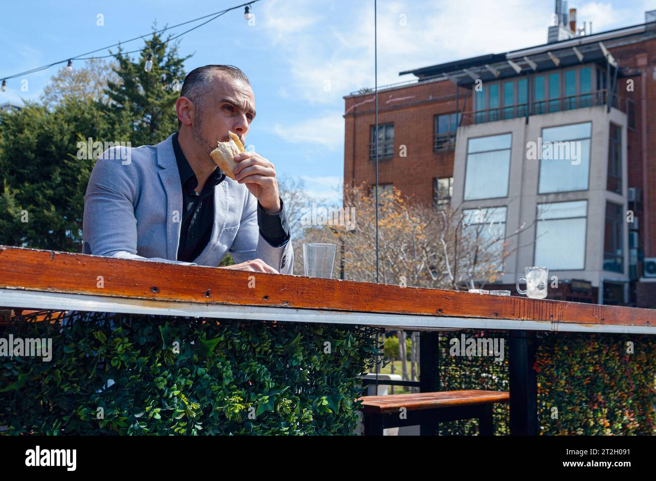 Uomo d'affari caucasico adulto, indossa una giacca, beve un panino a colazione, mangia e guarda avanti e pensa. posti a sedere al bar all'esterno del ristorante Foto Stock