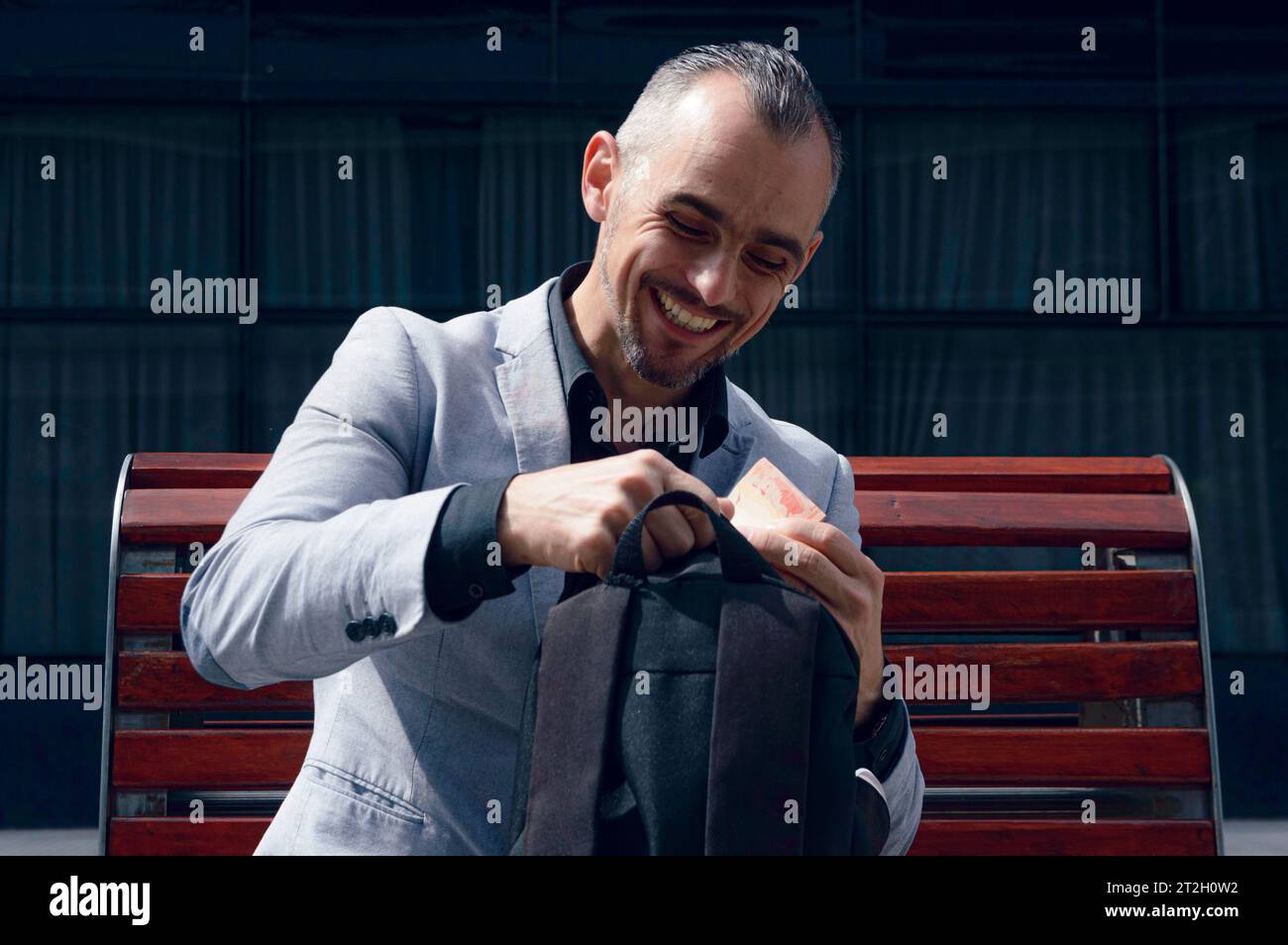 un uomo d'affari adulto, con i capelli corti, la barba e indossa un'elegante giacca grigia, è seduto all'aperto, ridendo felicemente perché ha i soldi nello zaino Foto Stock
