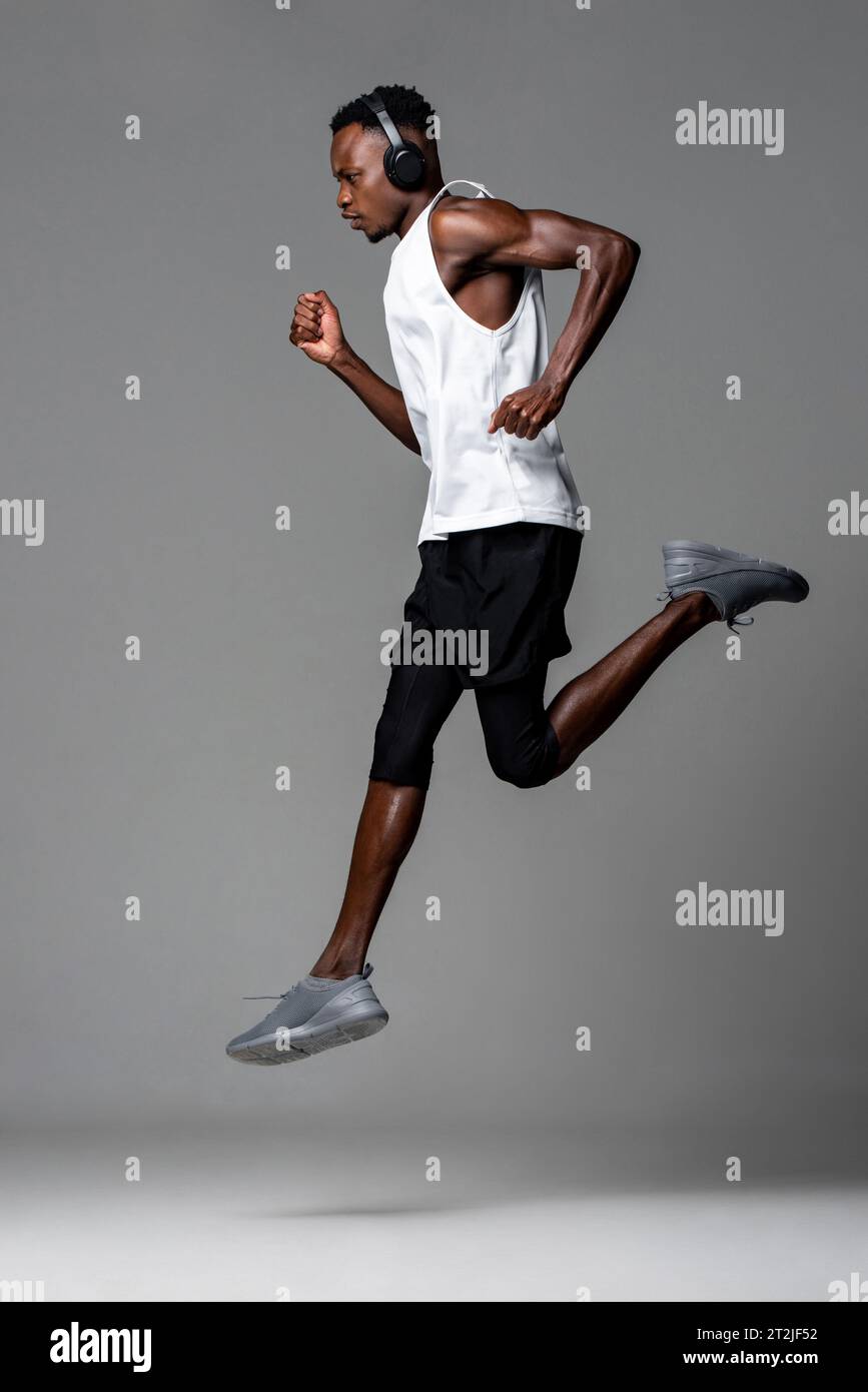 Sportivo africano muscoloso a tutta lunghezza con abbigliamento sportivo e cuffie che ascolta musica e corre durante l'allenamento su sfondo grigio Foto Stock