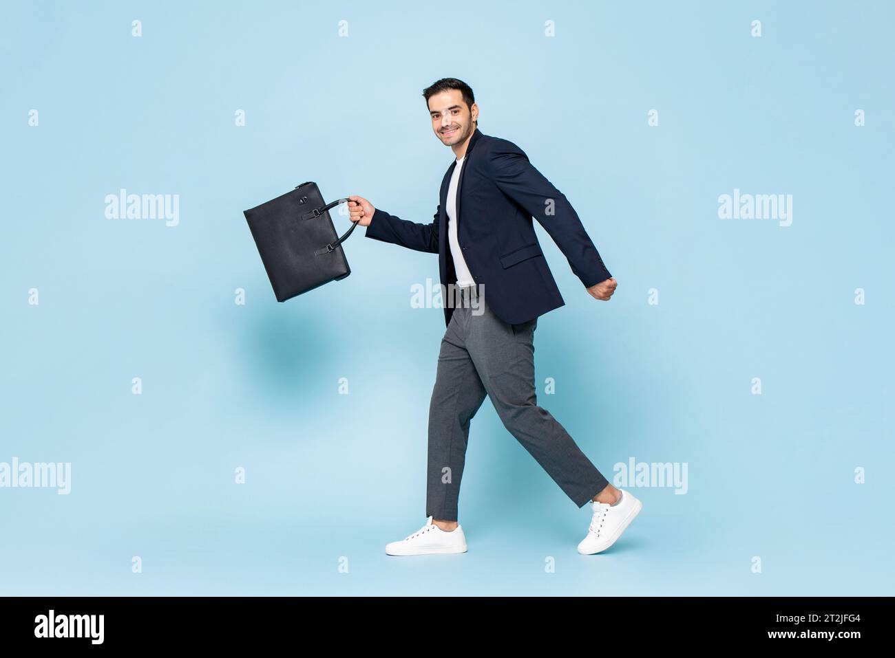 Imprenditore maschio con tutto il corpo in abiti eleganti e casual, braccia oscillanti con valigetta e guarda la macchina fotografica con sorriso mentre ti sposti per andare al lavoro Foto Stock