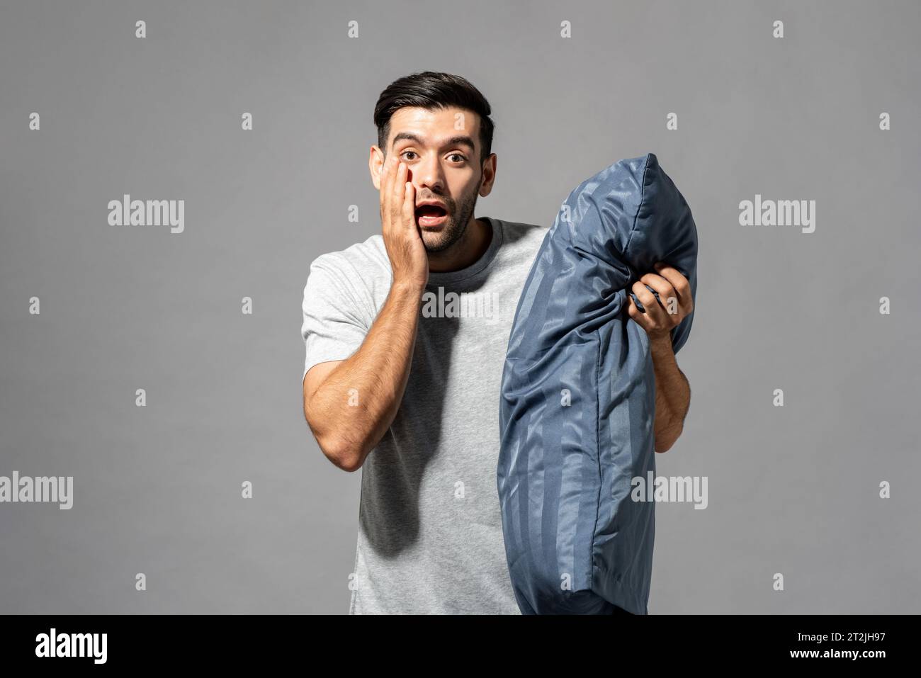 Sorpreso uomo caucasico con un morbido cuscino che tocca la guancia e guarda la fotocamera con la bocca aperta su sfondo grigio dello studio Foto Stock