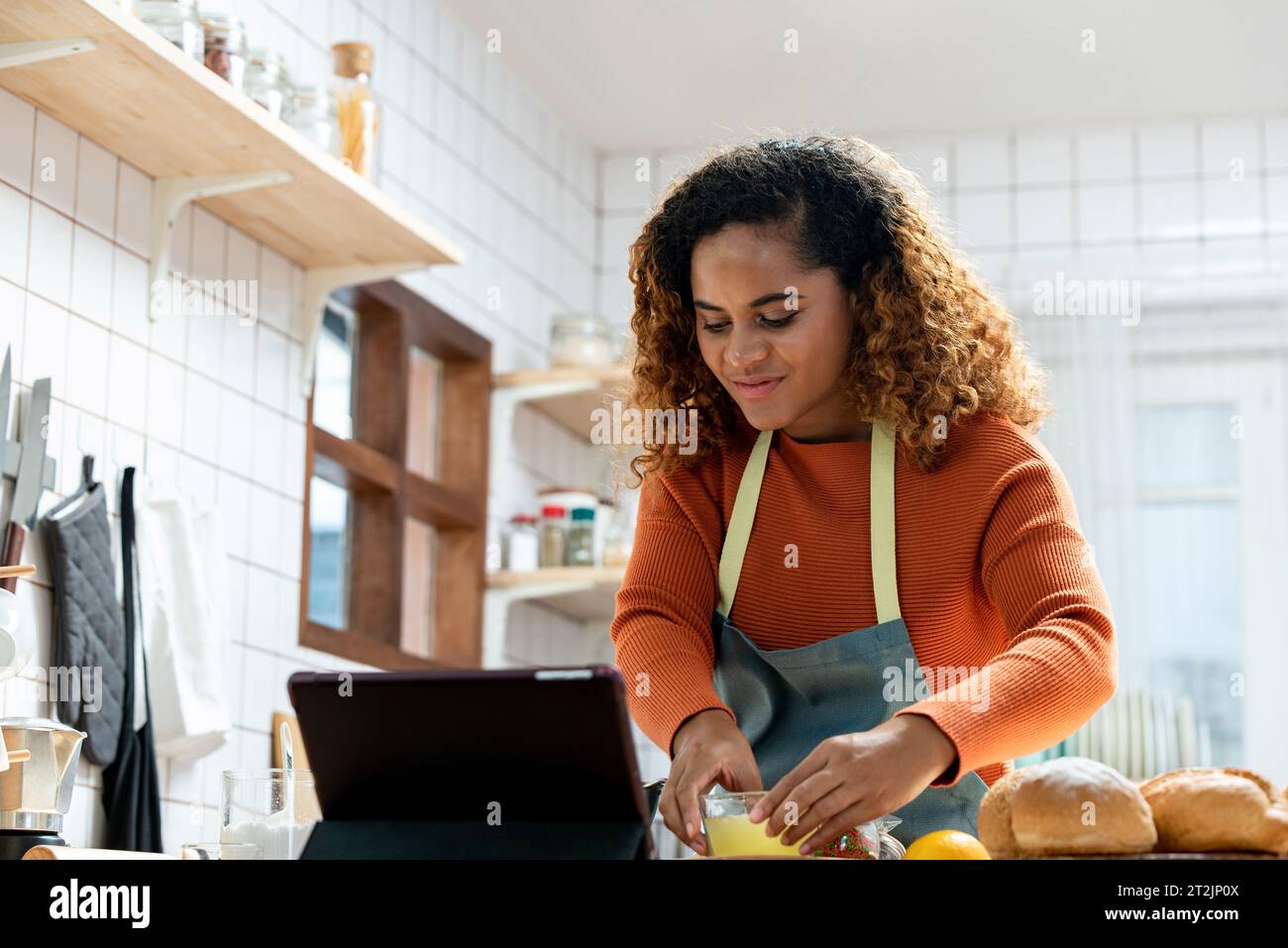 Donna afroamericana concentrata che prepara il cibo a tavola in cucina mentre registra il processo su tablet per vlog Foto Stock