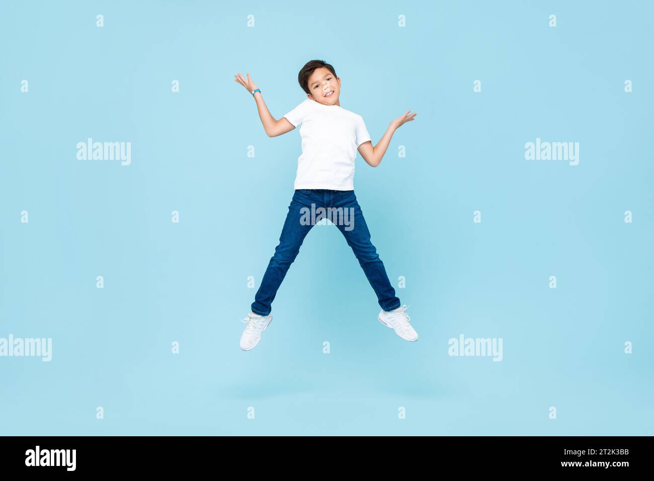 Simpatico ragazzo asiatico misto che salta e stringe le spalle in uno studio isolato di colore blu chiaro Foto Stock