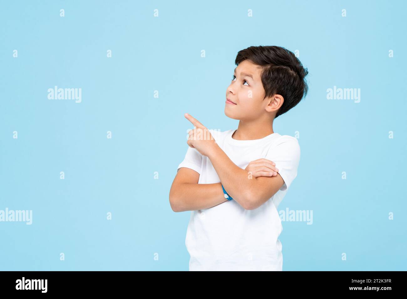 Simpatico ragazzo sorridente con una t-shirt bianca semplice che guarda e punta la mano verso l'alto su uno sfondo isolato di colore blu chiaro da studio Foto Stock