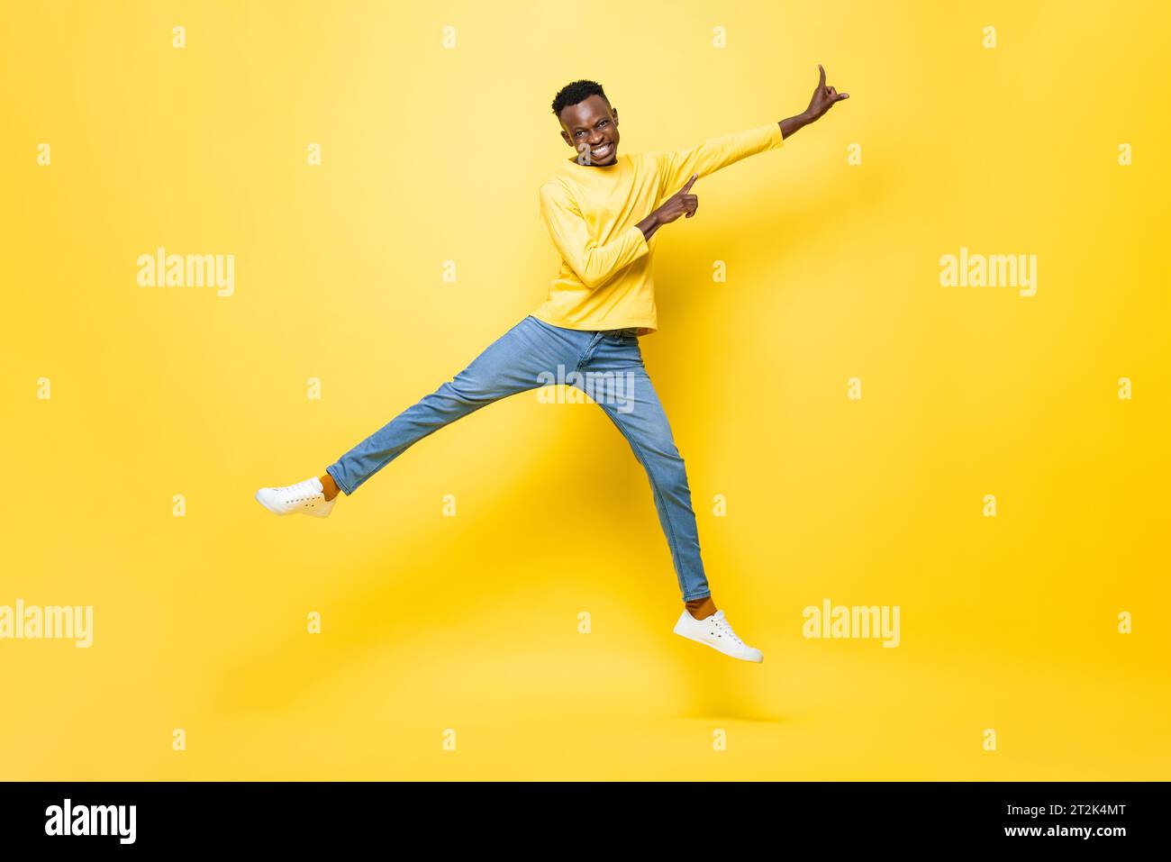 Ritratto a lunghezza intera di un giovane africano sorridente che salta e punta le mani su uno sfondo isolato giallo studio Foto Stock