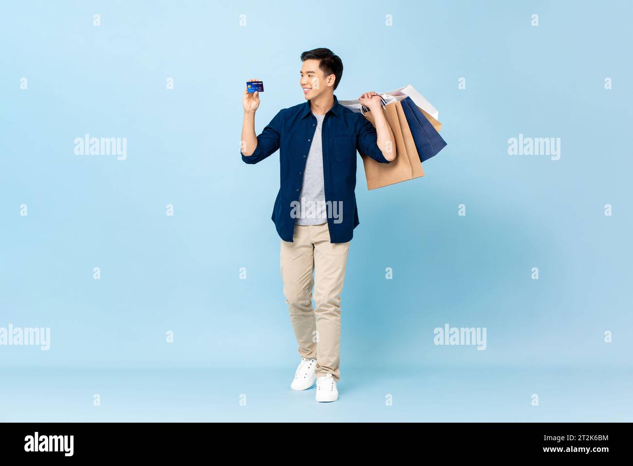 Ritratto a figura intera di un affascinante turista asiatico che porta con sé borse per la spesa e tiene una carta di credito in uno studio isolato di colore blu chiaro Foto Stock