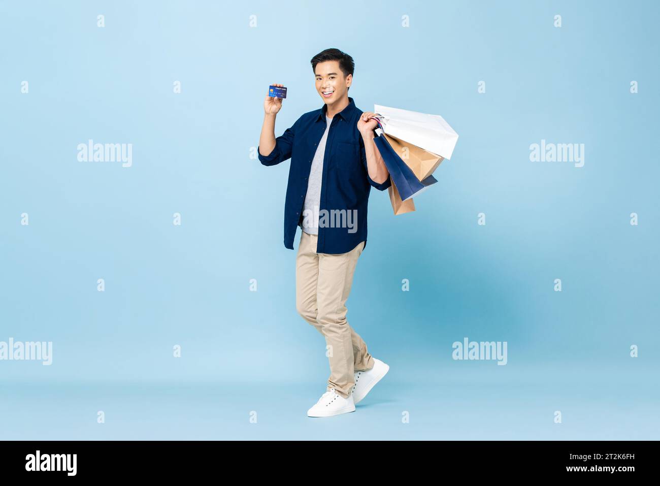 Ritratto a figura intera di un affascinante turista asiatico che porta con sé borse per la spesa e tiene una carta di credito in uno studio isolato di colore blu chiaro Foto Stock