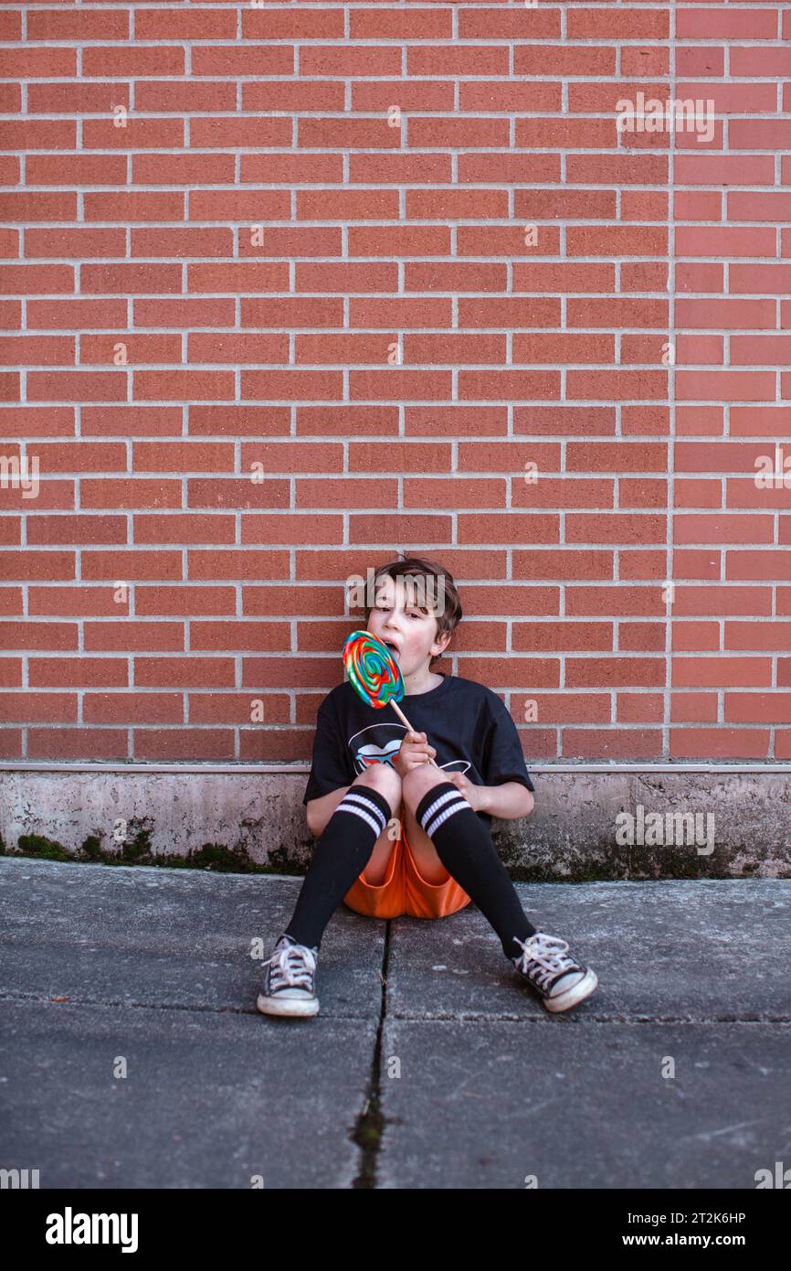 Young Boy Licking Giant Rainbow Lollipop di Brick Wall Foto Stock