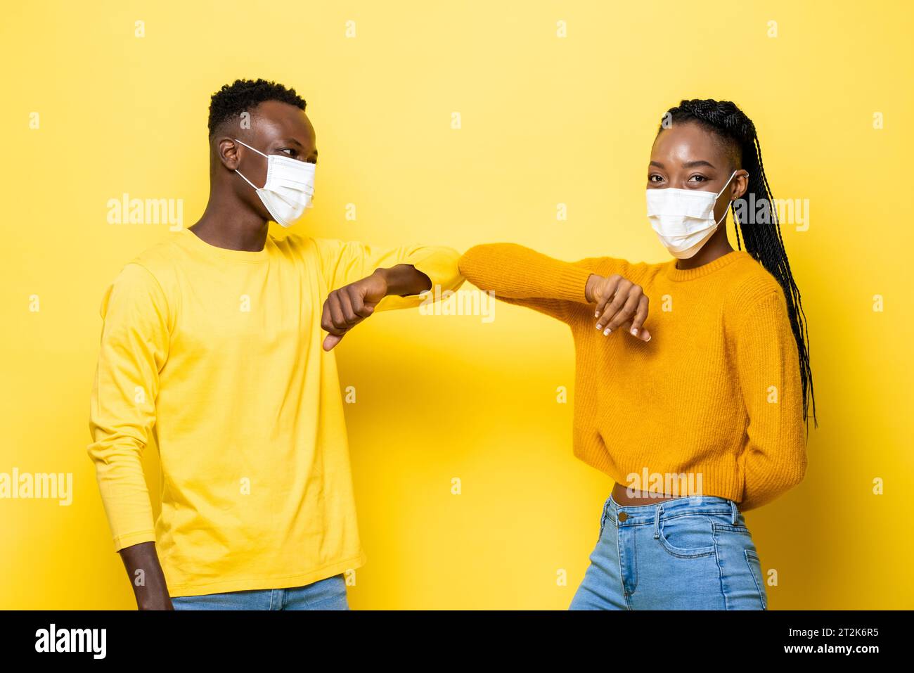 Giovane coppia afroamericana indossando maschere e urtare i gomiti per il saluto durante la pandemia COVID-19 in uno sfondo giallo isolato studio Foto Stock