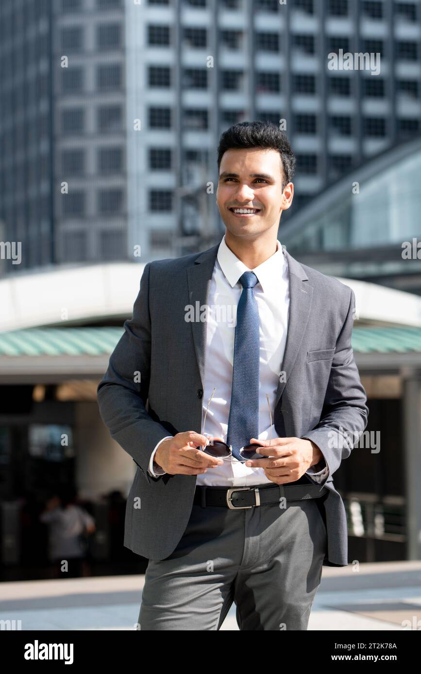 Ritratto di un elegante e sorridente uomo d'affari indiano che indossa una tuta grigia e regge gli occhiali da sole mentre si trova in piedi contro gli edifici della città Foto Stock