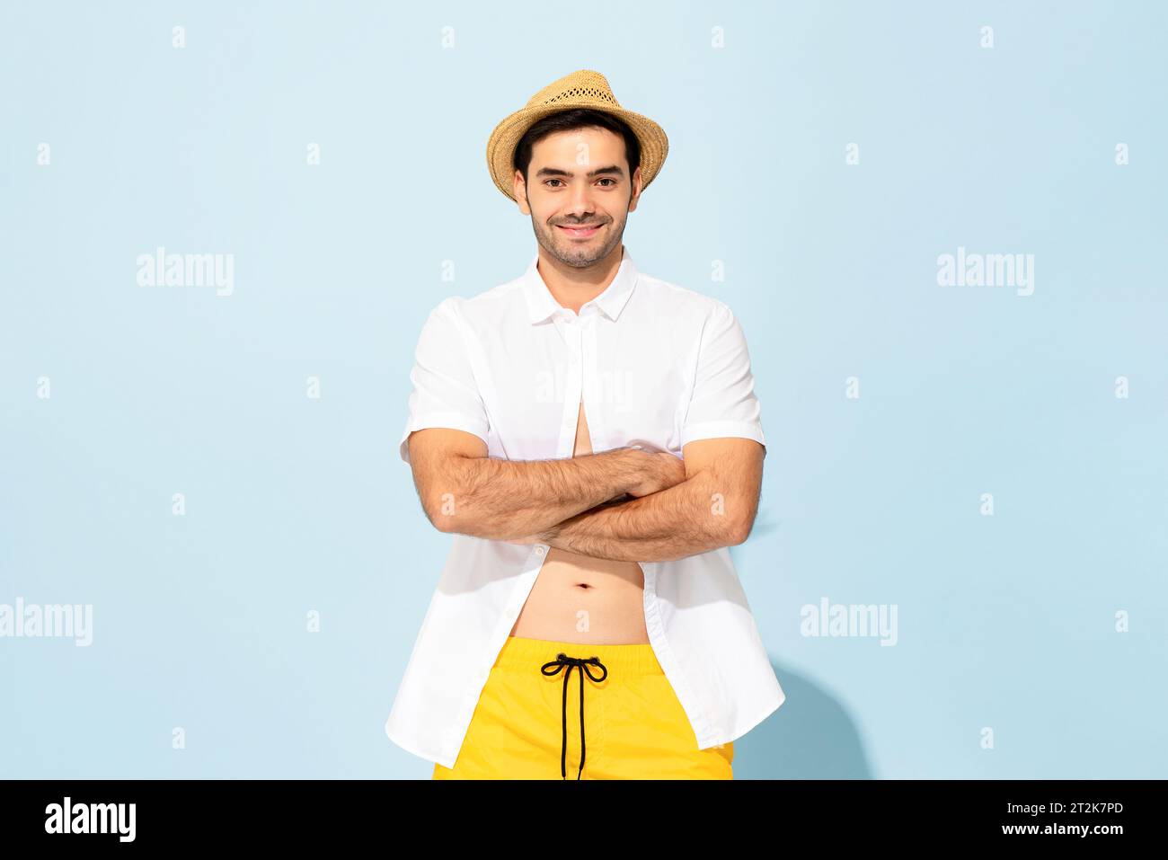 Bell'uomo caucasico in outfit estivo con cappello sorridente e braccia incrociate su sfondo blu chiaro isolato in studio Foto Stock