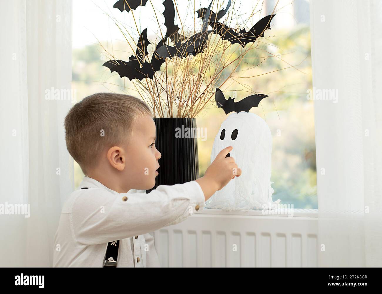 Concetto di Halloween. Niente faccia. Le mani del bambino vicino alla finestra, sullo sfondo di un vaso con rami asciutti e pipipistrelli di carta nera. Primo piano Foto Stock
