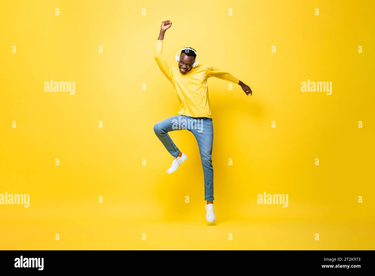 Ecstatico giovane africano che indossa delle cuffie per ascoltare musica e ballare con la mano in alto su uno sfondo isolato di colore giallo studio Foto Stock