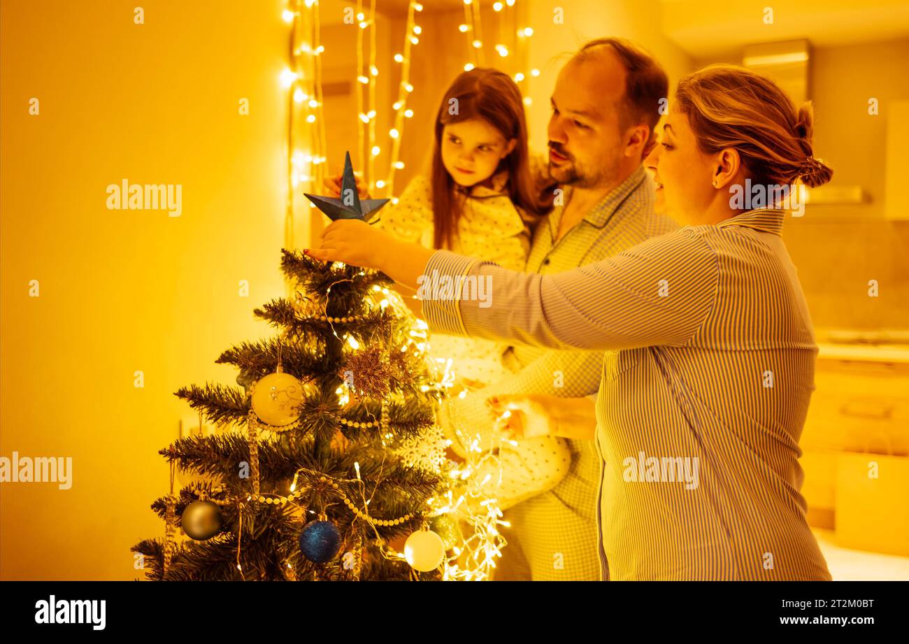 Mamma, papa' e piccola figlia decorano l'albero di Natale a casa. Un giovane uomo, una donna affascinante e il loro bambino carino si stanno preparando per il nuovo anno Foto Stock