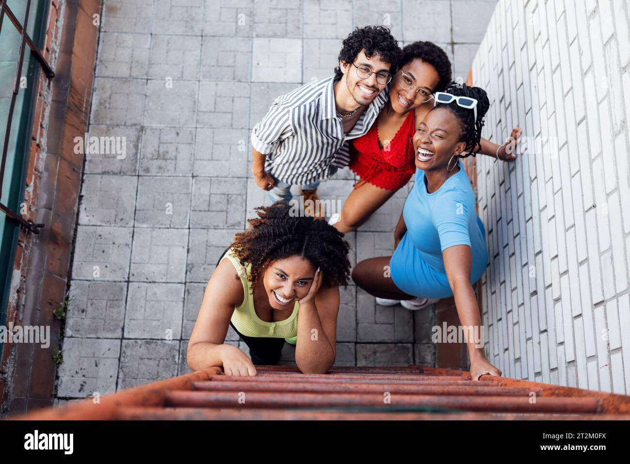 Tre bellissime donne africane e un uomo di razza mista sono in piedi vicino alle scale di ferro sulla casa sul tetto e sorridono. Gruppo di amici multietnici in abiti casual Foto Stock