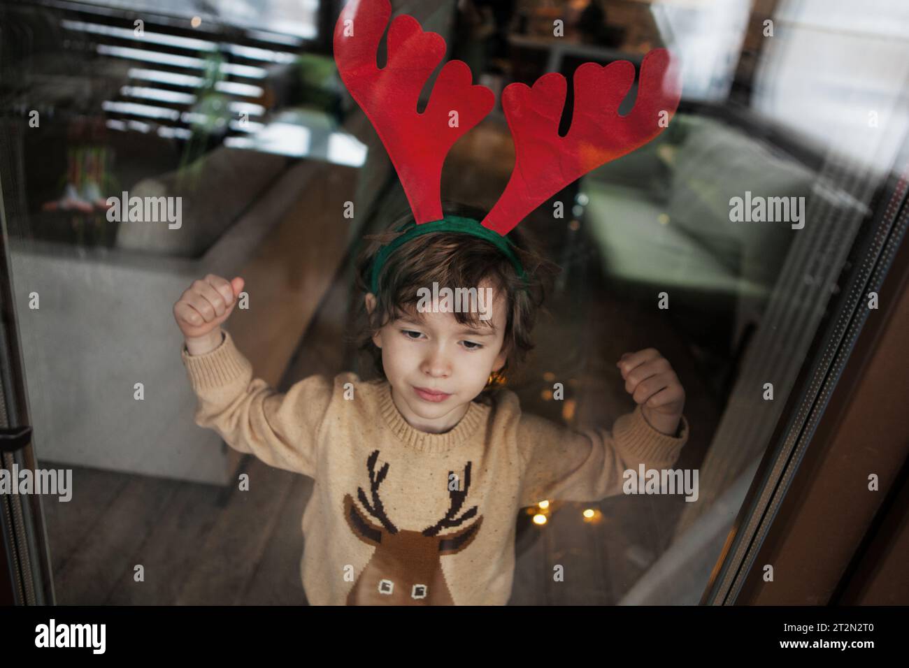 Il bambino con maglione a maglia e cervo sta vicino alla finestra, guarda all'esterno. Foto realistiche sullo stile di vita. Foto Stock