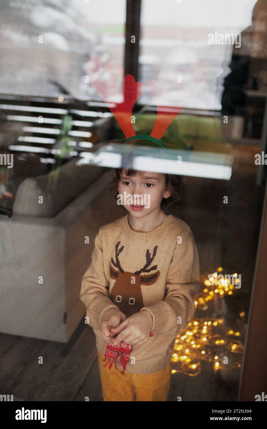 Bambino piccolo in maglione di cervo beige lavorato a maglia che indossa le corna delle renne a casa e guarda attraverso il vetro della finestra. Atmosfera natalizia. Foto Stock