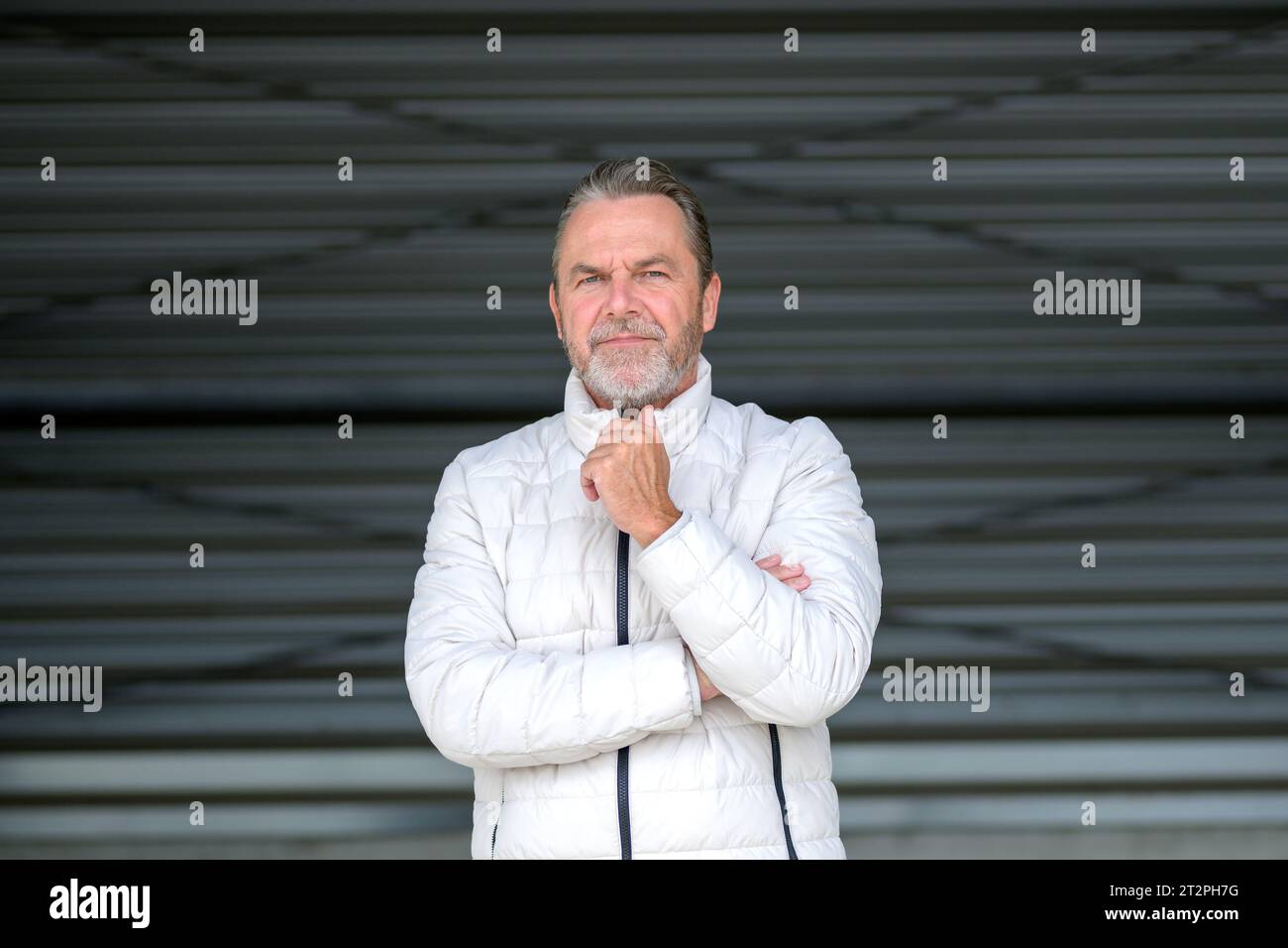 Attraente uomo dai capelli grigi che indossa una giacca invernale bianca con una mano sulla giacca che sembra amichevole con la fotocamera, davanti a una parete in ferro ondulato Foto Stock