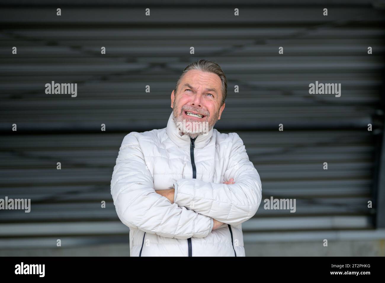 Attraente uomo dai capelli grigi con una giacca invernale bianca che guarda brividi al cielo di fronte a una parete in ferro ondulato Foto Stock