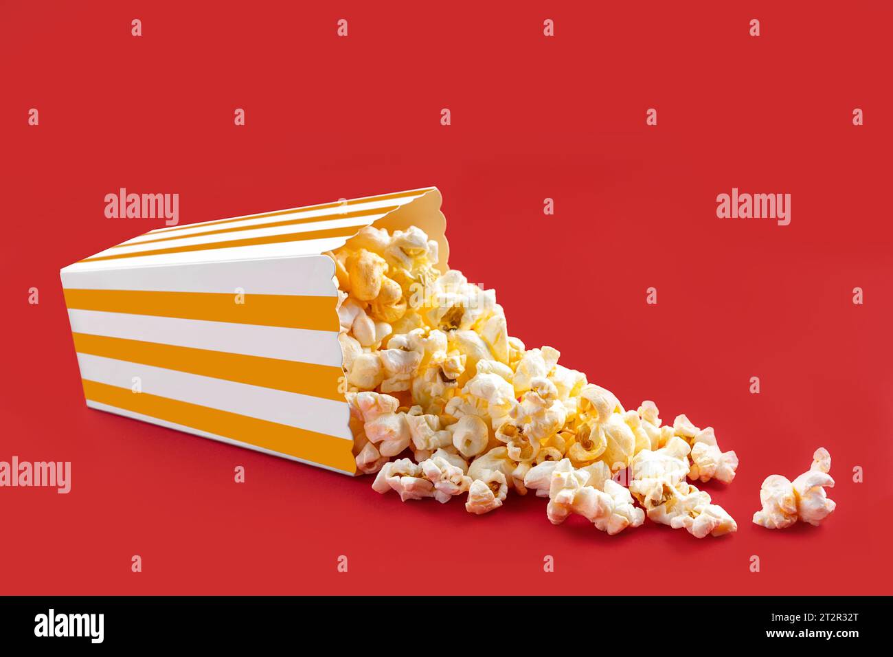 Gustosi popcorn al formaggio che cadono da una scatola di cartone a righe gialle o da un secchio, isolato su sfondo rosso. Dispersione di semi di popcorn. Fast food, snack. Foto Stock