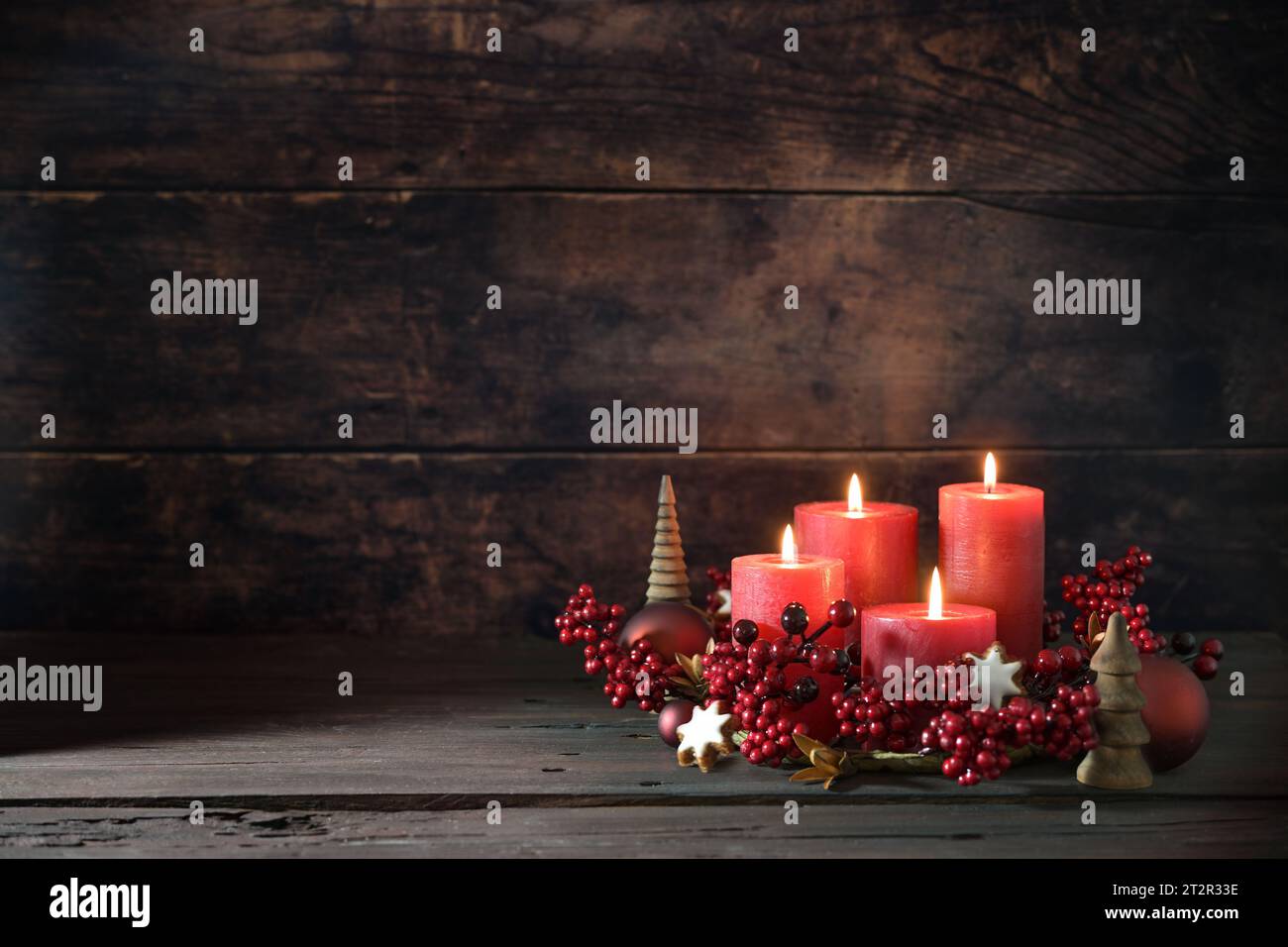 Quarto avvento con candele rosse accese in una corona di frutti di bosco decorativi, palle di Natale e biscotti con stelle di cannella su uno sfondo rustico scuro, Foto Stock