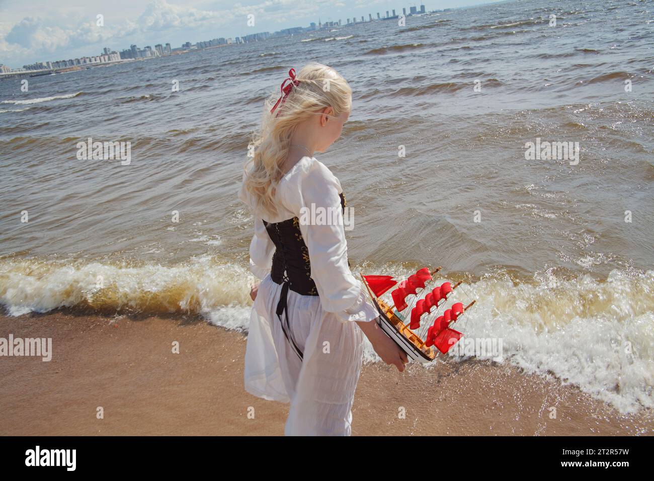 Adorabile giovane donna che cammina all'aperto. Bella signora in piedi sulla spiaggia di mare con un piccolo yacht a vela rossa Foto Stock