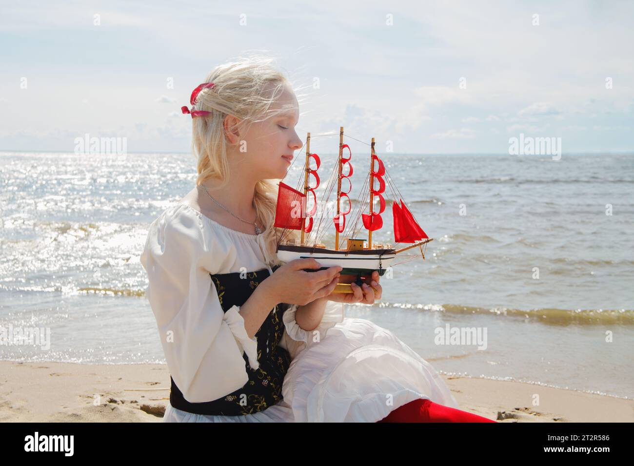 Bella giovane donna che regge un piccolo yacht con vele rosse sulla spiaggia di mare Foto Stock