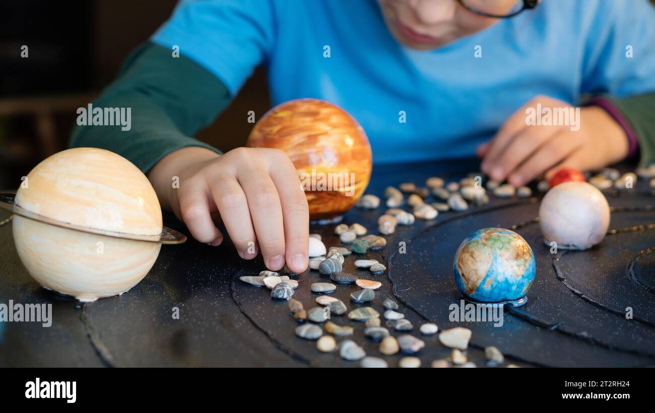 Primo piano delle mani di un bambino che posiziona pianeti dipinti e ciottoli su una superficie scura, reinventando un colorato modello di sistema solare Foto Stock