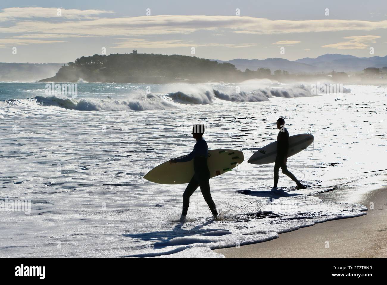 Paesaggio autunnale con due surfisti che camminano nel surf portando tavole da surf retroilluminate con la Penisola Maddalena Sardinero Santander Cantabria Spagna Foto Stock