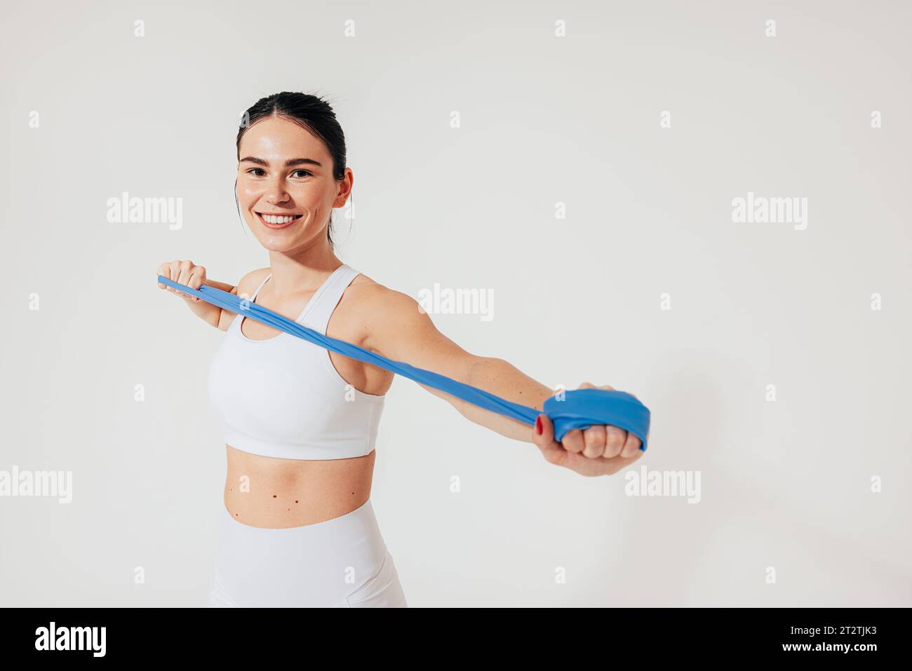 Una donna allegra in abbigliamento da fitness bianco si sta scaldando le braccia con una fascia di resistenza in studio Foto Stock