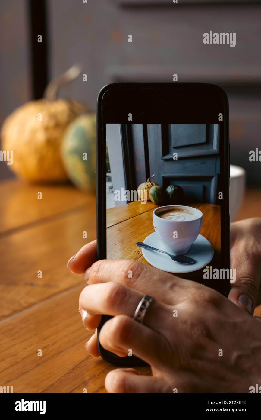 Donna che fa una foto al caffè mattutino. Tazza di caffè con zucche sullo sfondo sullo schermo del telefono. Telefono cellulare in mano donna. Concetto di blog. Foto Stock