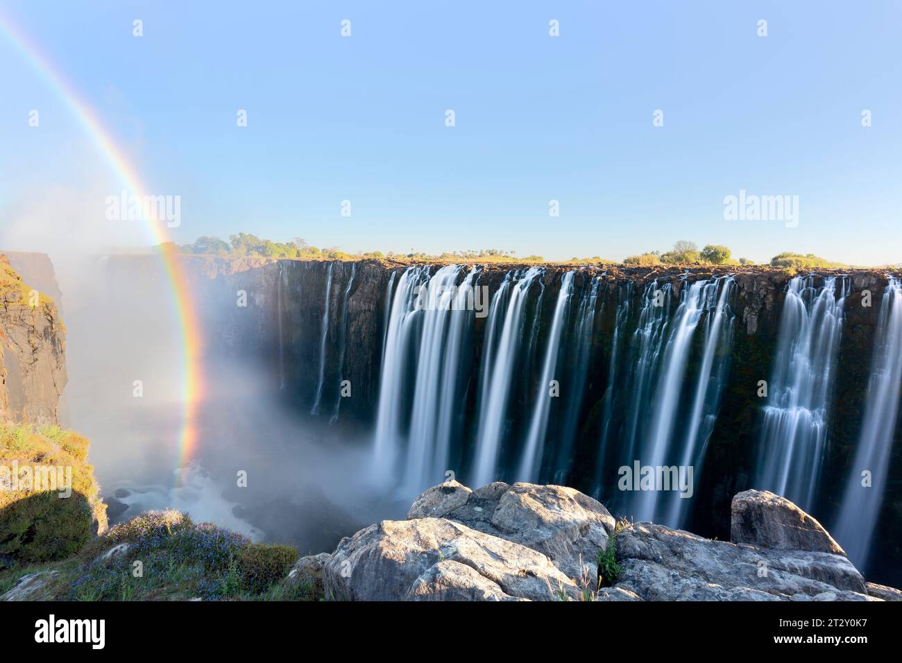 Foto panoramica della cascata delle Cascate Vittoria sul fiume Zambesi a flusso molto alto in tarda serata con spruzzi che si alzano e un intenso arcobaleno sulle cascate Foto Stock