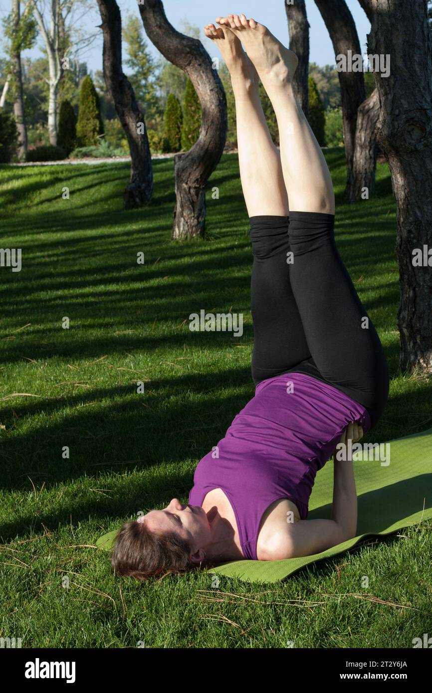 una donna di mezza età pratica yoga all'aperto nel parco cittadino. gli yogini principianti fanno asana invertita, sdraiati sul tappetino, le gambe in alto, appoggiandosi sulle spalle. Fitness, str Foto Stock