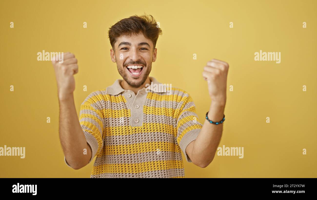 Un giovane arabo sicuro di sé che apprezza la pura felicità, festeggia con gioia la sua vittoria con un sorriso luminoso, in piedi contro un vibrante e isolato ba gialla Foto Stock