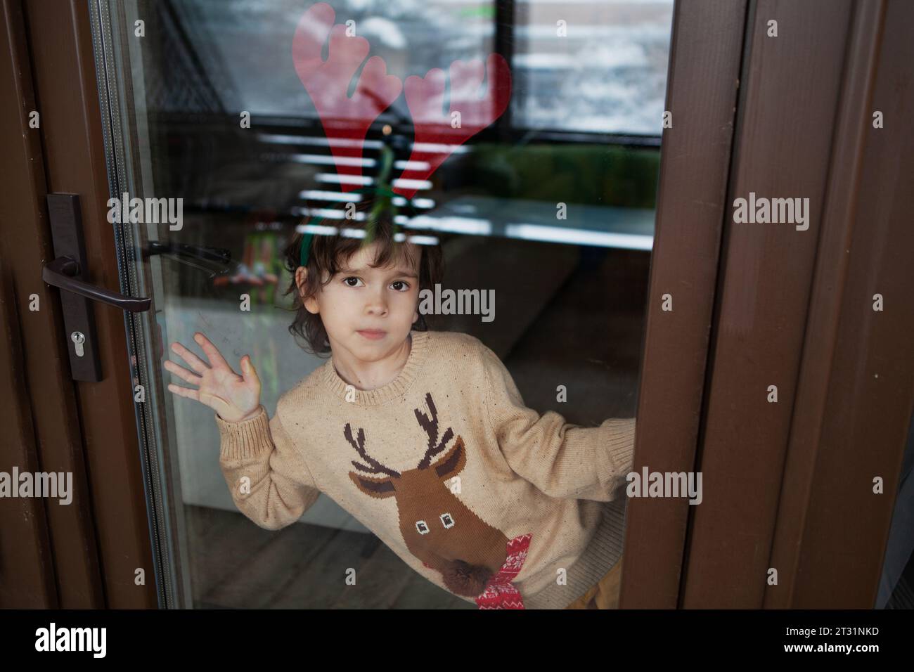 Adorabile bambino in maglione di cervo beige lavorato a maglia che indossa le corna delle renne a casa e guarda la macchina fotografica attraverso il vetro della finestra. Foto Stock