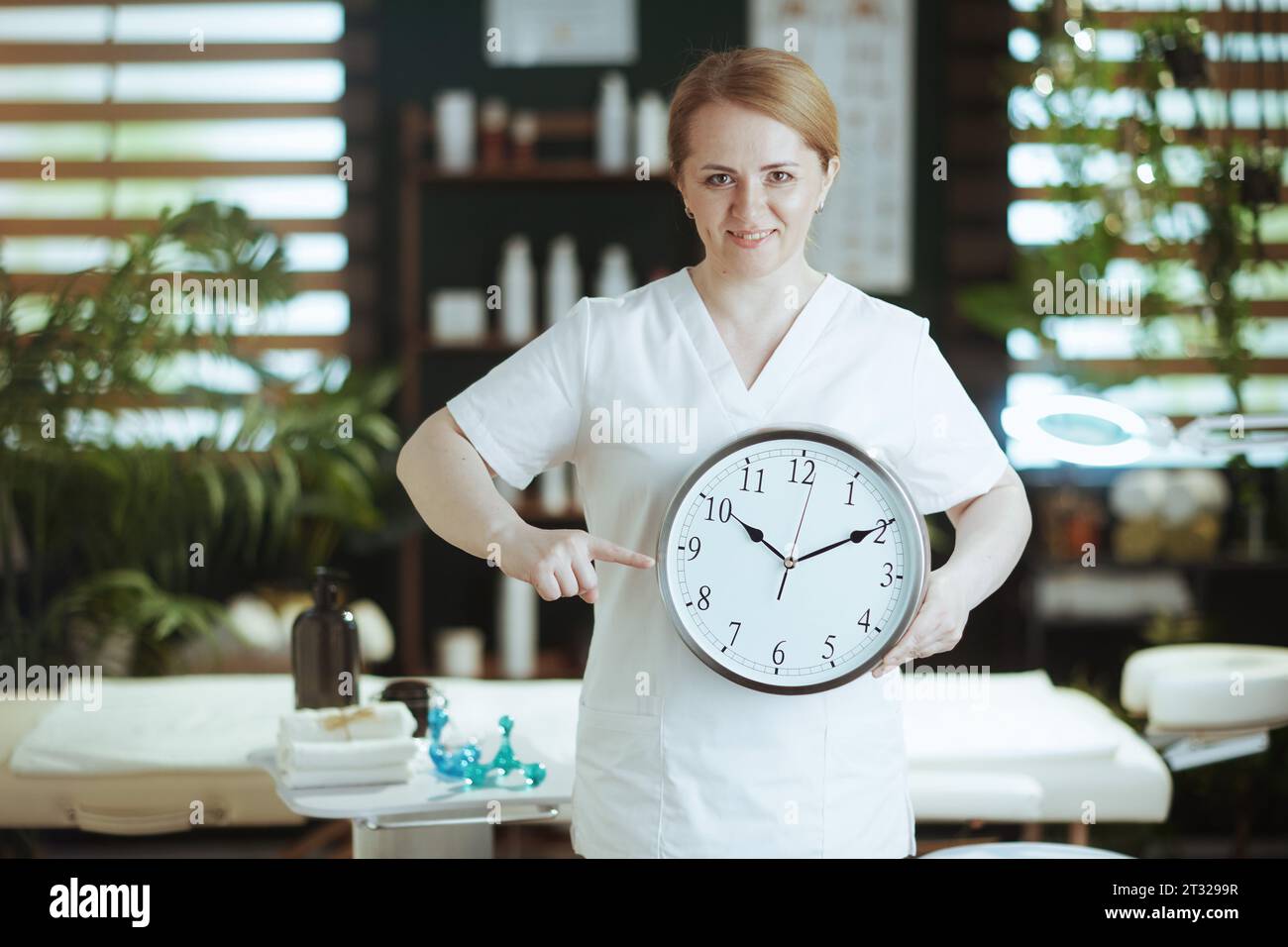 Il tempo dell'assistenza sanitaria. terapista di massaggio medico femminile sorridente in cabina di massaggio con orologio. Foto Stock