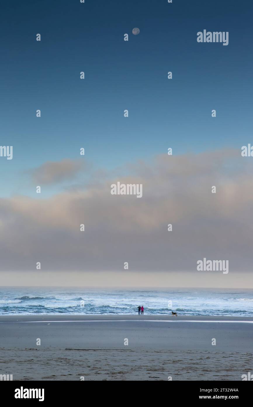 Due persone con il loro cane su una spiaggia sabbiosa che guarda la luna piena sull'Oceano Pacifico a Neskowin, Oregon Foto Stock